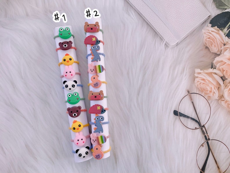 May include: Two white cylindrical displays labeled #1 and #2, each holding a vertical stack of colorful elastic hair ties. The hair ties feature various small, flat, cartoon-style animal and creature charms, including frogs, bears, chicks, bunnies, pandas, dinosaurs, and hedgehogs.