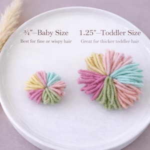 K&ouml;nnte beinhalten: Zwei runde, mehrfarbige Garn-Pompon-Haaraccessoires in Pastellt&ouml;nen wie Gelb, Rosa, Gr&uuml;n und Blau. Das kleinere ist mit 3/4 inch f&uuml;r Babys und das gr&ouml;&szlig;ere mit 1.25 inch f&uuml;r Kleinkinder beschriftet, beide liegen auf einem wei&szlig;en Teller.