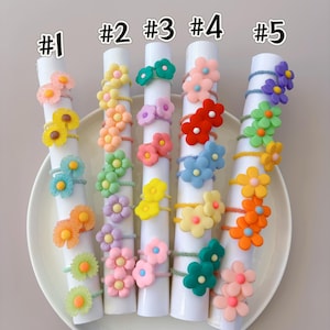May include: Five white cylindrical displays labeled #1 through #5, each holding multiple elastic hair ties adorned with colorful, multi-petaled flower charms in various shades of pink, yellow, orange, green, blue, purple, and red.
