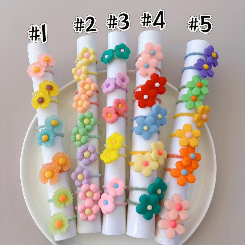 May include: Five white cylindrical displays labeled #1 through #5, each holding multiple elastic hair ties adorned with colorful, multi-petaled flower charms in various shades of pink, yellow, orange, green, blue, purple, and red.