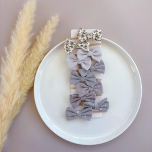 May include: A set of eight hair bows in various shades of gray and white with floral and polka dot patterns. The bows are attached to alligator clips.