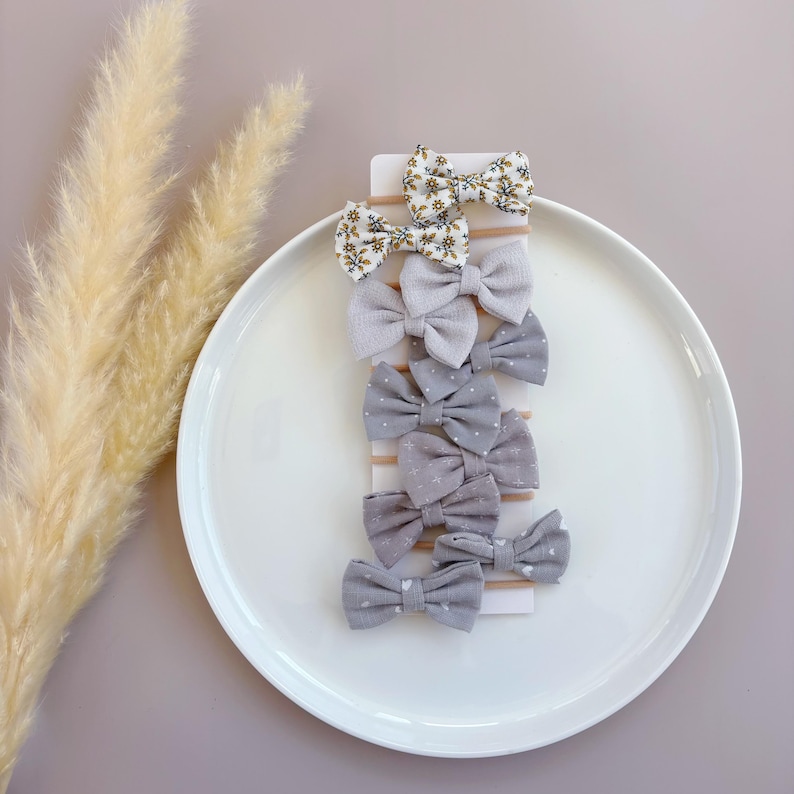 May include: A set of eight hair bows in various shades of gray and white with floral and polka dot patterns. The bows are attached to alligator clips.