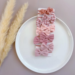 May include: A set of 8 pink and white hair bows with different patterns, including gingham, polka dots, and floral prints. The bows are arranged on a white card with a pink background.