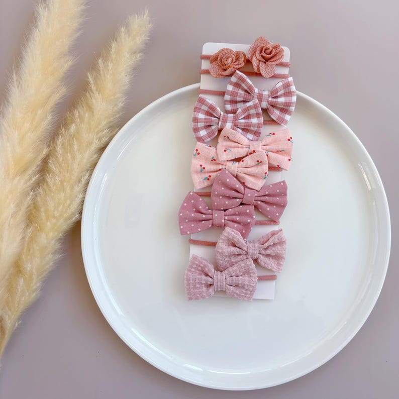 May include: A set of 8 pink and white hair bows with different patterns, including gingham, polka dots, and floral prints. The bows are arranged on a white card with a pink background.
