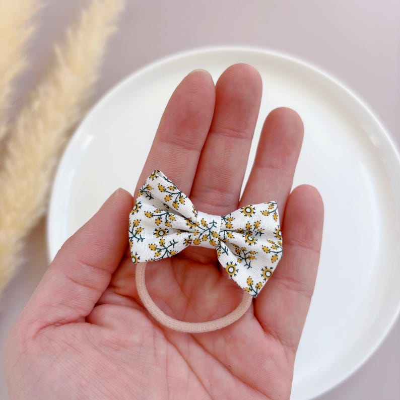 May include: A white fabric hair bow with a yellow floral pattern. The bow is tied around a nude elastic hair tie.