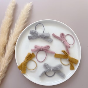 May include: Six fuzzy hair ties with bows in various colors, including gray, pink, and yellow. Each bow has a small pearl accent in the center.