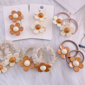 May include: A collection of hair accessories featuring daisy designs. The set includes hair clips, scrunchies, and hair ties. The flower petals are a mix of white and brown, with a cream-colored center. The accessories are arranged on a white surface.