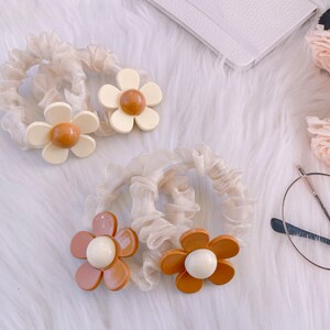 May include: Four ivory scrunchies with daisy flower accents in shades of cream, brown, and white. The scrunchies are made of a sheer, ruffled fabric. The background includes a white notebook, glasses, and roses.