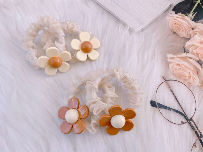 May include: Four ivory scrunchies with daisy flower accents in shades of cream, brown, and white. The scrunchies are made of a sheer, ruffled fabric. The background includes a white notebook, glasses, and roses.