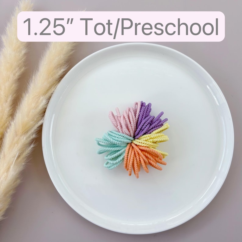 Op de afbeelding: Een set van 3,18 cm gekleurde haarbandjes in de vorm van een bloem. De haarbandjes zijn in tinten roze, paars, geel en groen. Tot/Preschool.