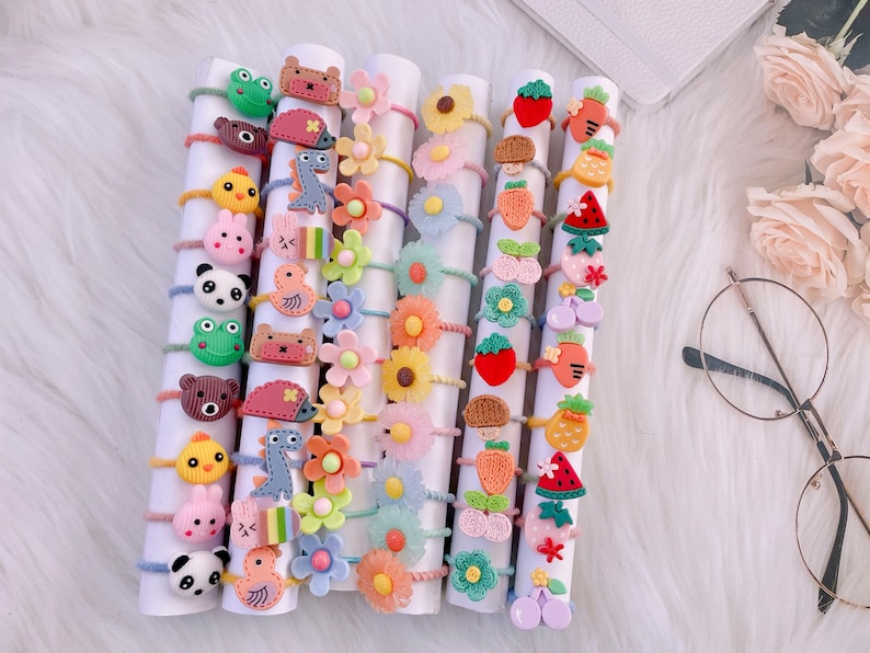 May include: A collection of colorful hair ties featuring various decorative charms including animals, flowers, fruits, and vegetables. The charms are arranged on white cylindrical holders, showcasing designs like pandas, frogs, bears, daisies, strawberries, and watermelons in a variety of bright colors.