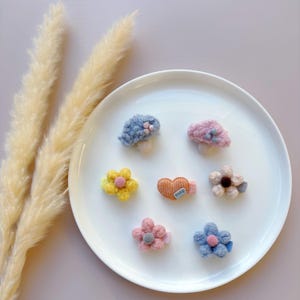 Pode incluir: Um conjunto de sete prendedores de cabelo felpudos coloridos. Os prendedores têm o formato de flores, corações e cogumelos. Os prendedores são de diferentes cores, incluindo rosa, azul, amarelo e marrom.