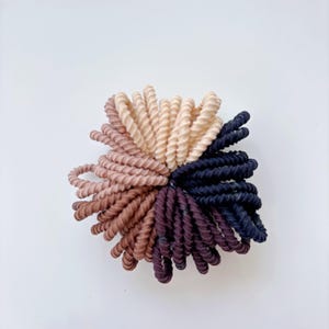 May include: A close-up of a set of hair ties in a variety of colors, including beige, brown, and black. The hair ties are arranged in a circular pattern, with the beige hair ties in the center and the brown and black hair ties surrounding them.