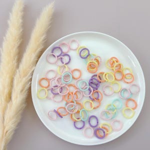 Op de afbeelding: Een wit bord gevuld met een verscheidenheid aan kleurrijke haar elastiekjes. De haar elastiekjes zijn gemaakt van een zacht, rekbaar materiaal en zijn verkrijgbaar in een reeks pastelkleuren, waaronder roze, paars, blauw en geel.