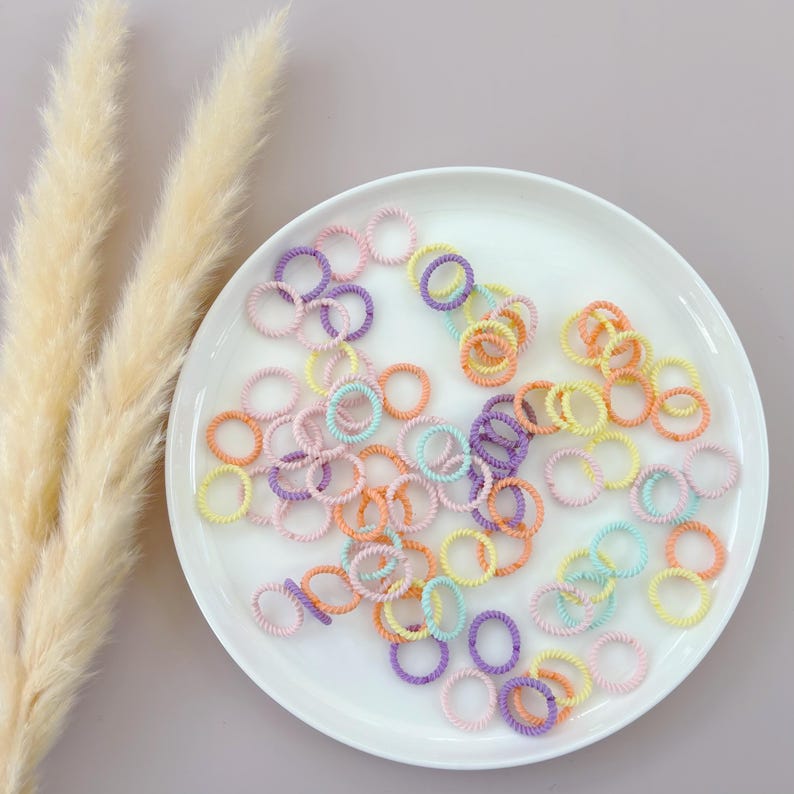 Op de afbeelding: Een wit bord gevuld met een verscheidenheid aan kleurrijke haar elastiekjes. De haar elastiekjes zijn gemaakt van een zacht, rekbaar materiaal en zijn verkrijgbaar in een reeks pastelkleuren, waaronder roze, paars, blauw en geel.