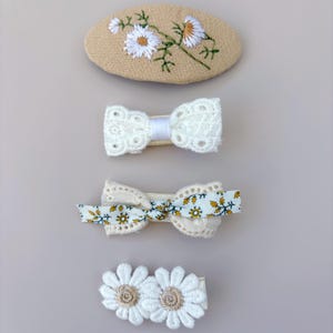 May include: A set of four hair clips including an oval beige clip with embroidered white daisies, a white lace bow clip, a cream lace bow clip layered with a yellow and white floral ribbon, and a clip featuring two white daisy-shaped fabric flowers.