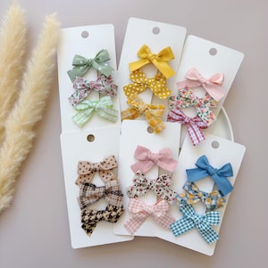 May include: A set of 12 hair bows in various colors and patterns, including floral, plaid, polka dot, and solid colors. The bows are displayed on white cards with a hole at the top for hanging.