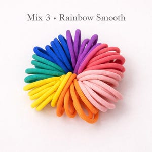 K&ouml;nnte beinhalten: Eine Ansammlung von elastischen Haargummis, die in einem kreisf&ouml;rmigen Muster angeordnet sind und ein Farbspektrum aus Blau, Gr&uuml;n, Gelb, Orange, Rosa und Violett aufweisen. Der Text 'Mix 3 &bull; Rainbow Smooth' steht &uuml;ber den Haargummis.