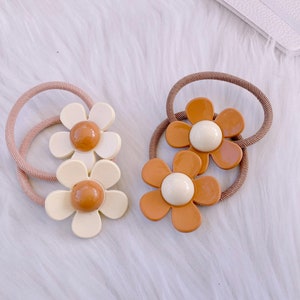 May include: Two daisy-shaped hair ties. One is cream-colored with a brown center, the other is brown with a cream center. Both have a brown elastic band. These hair accessories are perfect for adding a touch of floral flair to any hairstyle.