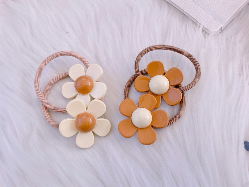 May include: Two daisy-shaped hair ties. One is cream-colored with a brown center, the other is brown with a cream center. Both have a brown elastic band. These hair accessories are perfect for adding a touch of floral flair to any hairstyle.