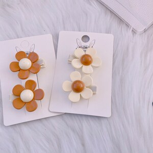 May include: Two sets of daisy-shaped hair clips. One set features brown petals with a white center, the other has off-white petals with a brown center. Each clip is attached to a small, light-colored barrette.