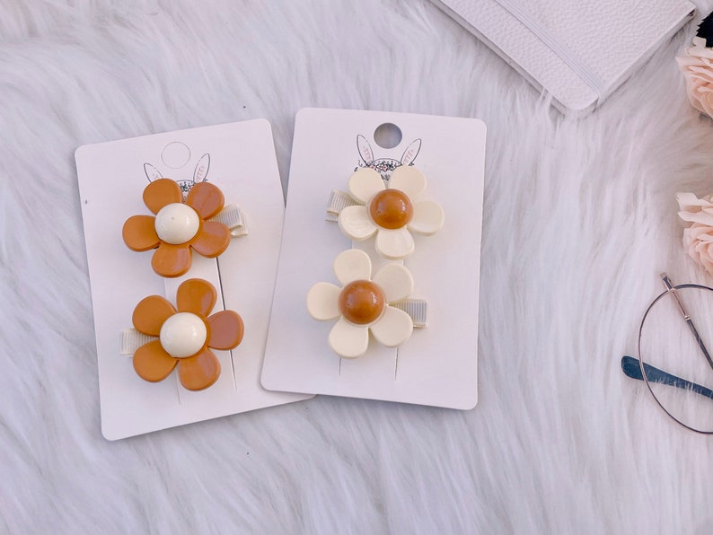 May include: Two sets of daisy-shaped hair clips. One set features brown petals with a white center, the other has off-white petals with a brown center. Each clip is attached to a small, light-colored barrette.