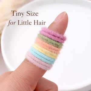 K&ouml;nnte beinhalten: Ein Stapel kleiner, flauschiger Haargummis in Pastellfarben wie Rosa, Gr&uuml;n, Pfirsich, Gelb und Blau. Der Text "Tiny Size for Little Hair" ist in Braun &uuml;ber den Haargummis aufgedruckt, die in einer vertikalen Reihe an einem Finger stecken.