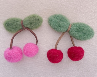 Puede incluir: Dos pinzas para el pelo de fieltro en forma de cereza. Una es verde y la otra roja. Ambas tienen cerezas de fieltro rosa.