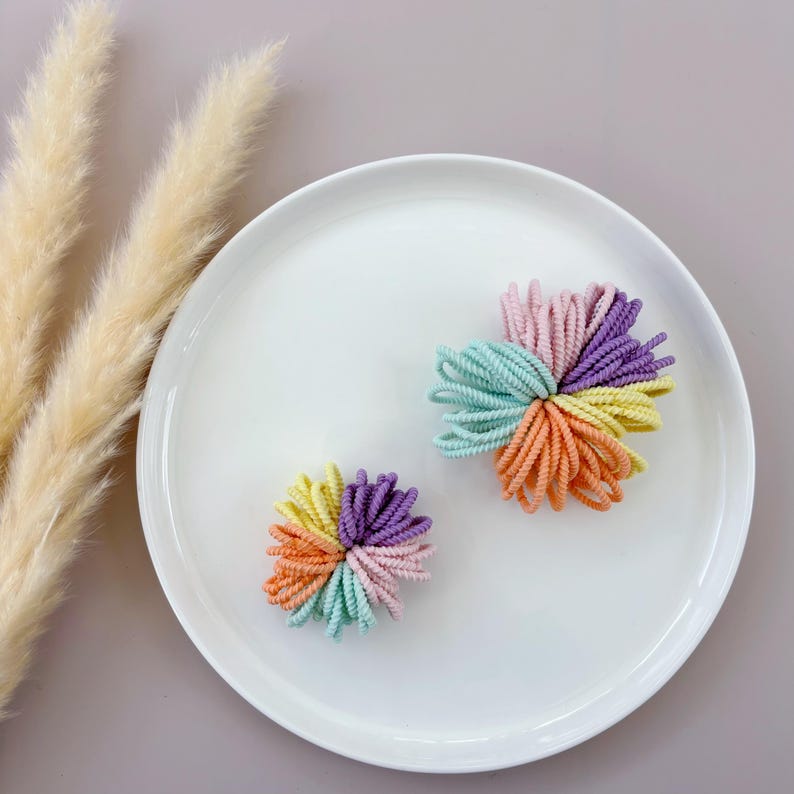 Op de afbeelding: Twee sets kleurrijke haar elastiekjes in een pastelkleurenschema. De haar elastiekjes zijn gerangschikt in een bloemvorm. De haar elastiekjes liggen op een wit bord.