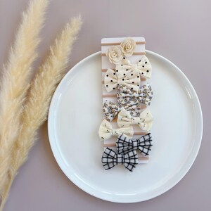 May include: Set of six hair bows with different patterns. The bows are made of white fabric with black and gold accents. The bows are attached to nude elastic hair ties.