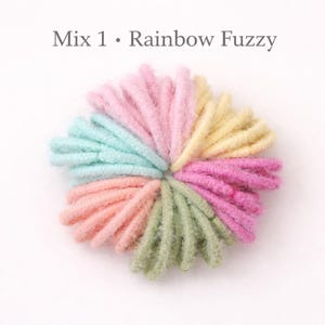 K&ouml;nnte beinhalten: Ein kreisf&ouml;rmiges B&uuml;ndel aus flauschigen Garnschlaufen in Pastellt&ouml;nen wie Rosa, Gelb, Hellblau, Pfirsich und Salbeigr&uuml;n. Der Text 'Mix 1 &bull; Rainbow Fuzzy' ist in Grau &uuml;ber dem Garnarrangement gedruckt, das eine weiche, strukturierte Optik aufweist.