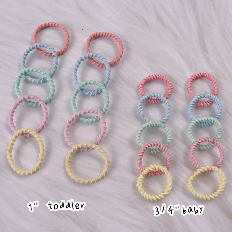 May include: Two sets of colorful hair ties, one set labeled "1" toddler and the other set labeled "3/4" baby. The hair ties are made of a braided rope material and come in pastel colors.