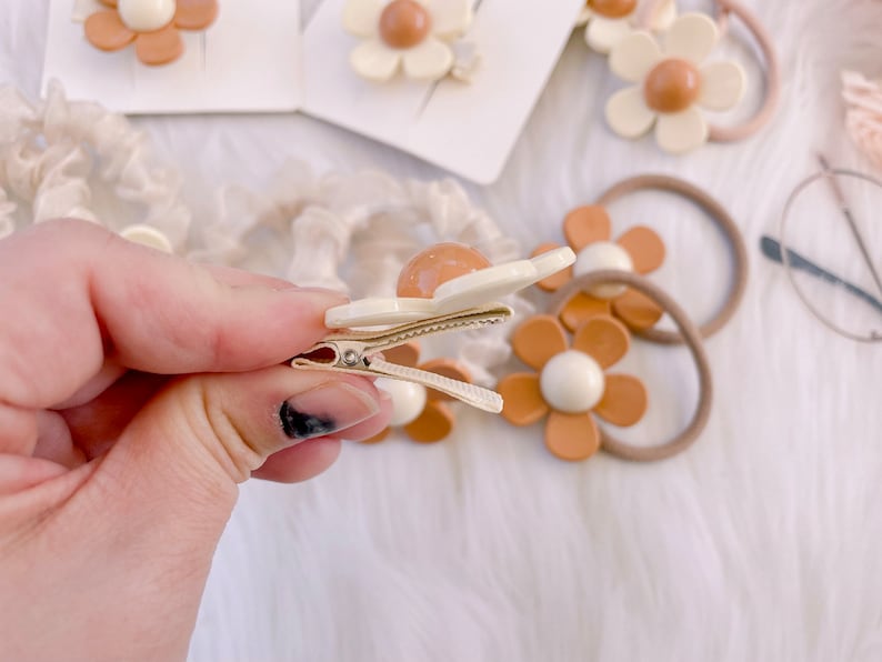 May include: A close-up of a daisy-shaped hair clip with a cream-colored flower and a tan center. The clip is held in a hand, with other hair accessories, including hair ties and more clips, visible in the background.