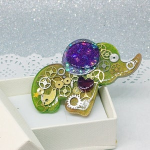 Puede incluir: Un broche de elefante verde y dorado con un corazón de purpurina morado y engranajes. El broche está en una caja blanca con una cinta blanca.