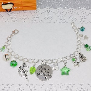 Könnte beinhalten: Silbernes Charm-Armband mit verschiedenen Anhängern, darunter eine Fee auf einem Mond, ein grüner Stern, ein Blatt und eine Glocke. Ein runder Anhänger lautet "Faith, Trust, & Pixie Dust". Das Armband hat auch grüne und klare Perlen.