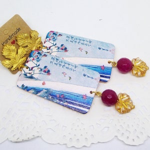 Könnte beinhalten: Ein Paar handgefertigte Ohrringe mit einem rechteckigen, blau-weißen Muster, goldenen Blattverzierungen und einem rosafarbenen Edelstein-Anhänger.