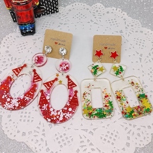 May include: Two pairs of handmade Christmas-themed earrings. The first pair features red and white party hats and snowflakes in a clear resin base. The second pair features green and gold Christmas trees and stars in a clear resin base. Both pairs have a small tag that says "Handmade with love".