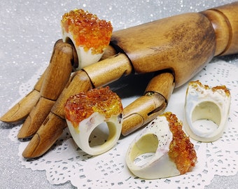 May include: Four white resin rings with orange and yellow crystal-like inclusions. The rings are displayed on a wooden hand.