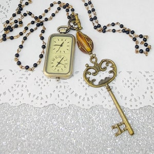 Puede incluir: Un collar con un reloj de bolsillo dorado y una cadena de cuentas negras. El reloj tiene una esfera blanca con marcadores de hora en números romanos negros y agujas negras. El collar también incluye un dije de llave decorativo y una pequeña cuenta de vidrio ámbar.