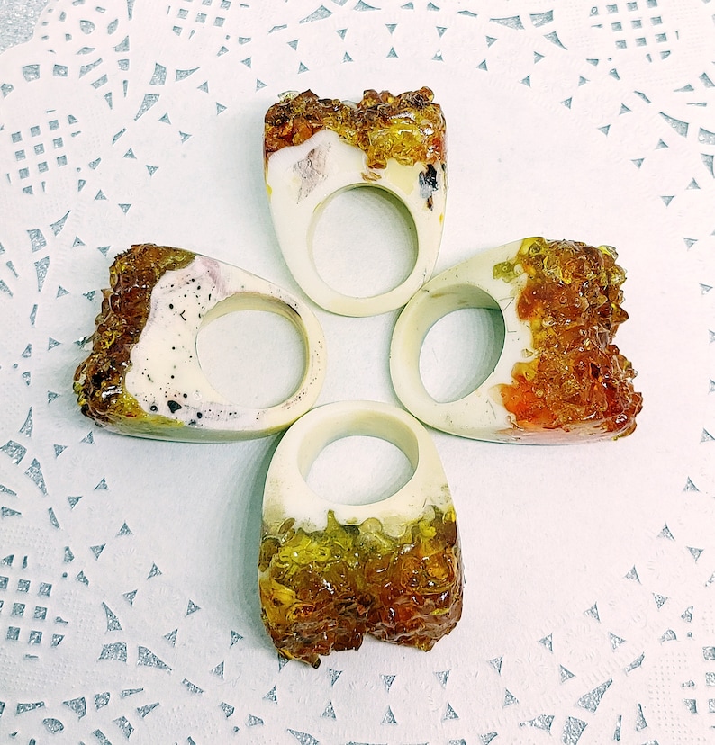 May include: Four resin rings with a white base and a clear resin top filled with amber-colored crystals. The rings are arranged in a cross shape.