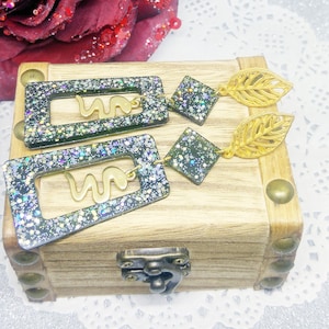 May include: A pair of dangle earrings with a black and green glitter resin base. The earrings feature a gold snake design and a gold leaf charm.