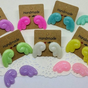 Puede incluir: Surtido de pendientes de alas de ángel con purpurina hechos a mano en varios colores, incluyendo rosa, verde, azul, amarillo, blanco y morado. Cada par se muestra en una pequeña tarjeta marrón con el texto "Handmade with Love".