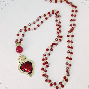 May include: A long necklace with a gold-toned heart-shaped pendant with a red centre. The necklace has a strand of small, red beads and a gold chain. The pendant is decorated with a gold-toned flame design.