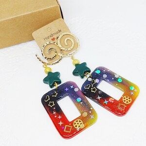 May include: Rainbow colored resin earrings with a gold swirl design and a green star charm. The earrings have a holographic finish and feature a variety of small details, including hearts, stars, and gears.