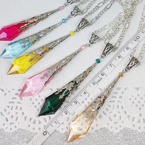 May include: Six silver necklaces with different colored crystal pendants. The pendants are shaped like teardrops and have intricate silver designs. The necklaces are laid out on a white surface with a ruler for scale.