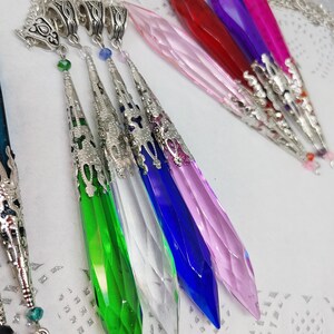 May include: A set of seven crystal pendants with silver-toned metal accents. The pendants are in various colors, including pink, red, purple, green, blue, and clear. The pendants are all shaped like a pointed teardrop.