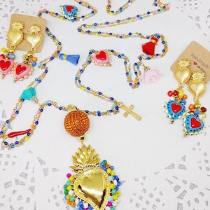 Puede incluir: Una colección de coloridas joyas de cuentas, que incluyen collares y pendientes. Los collares presentan colgantes de corazón, cruces y esferas naranjas. Los pendientes tienen diseños en forma de corazón. La joyería está sobre un fondo blanco.