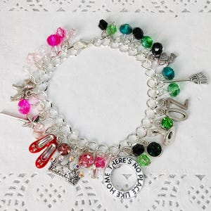Puede incluir: Un brazalete de plata con una variedad de dijes, que incluyen una corona, zapatillas de rubí, una escoba de bruja y un dije que dice "No hay lugar como el hogar". El brazalete también presenta cuentas de cristal rosa, verde y negro.