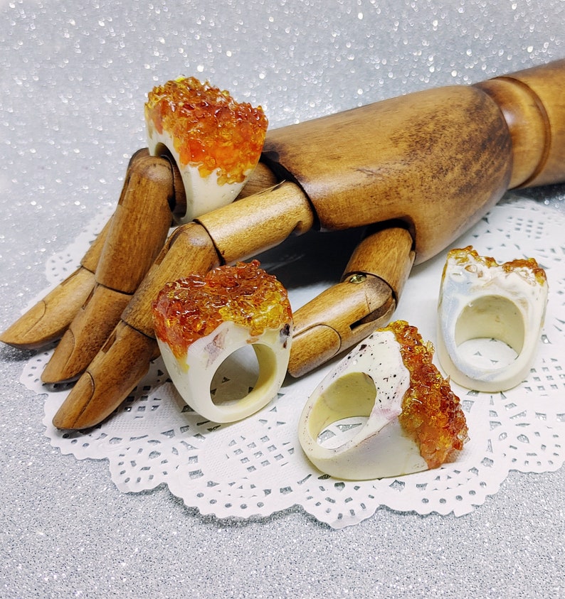 May include: Four white resin rings with orange and yellow crystal-like inclusions. The rings are displayed on a wooden hand.