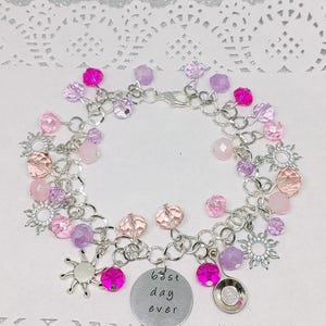 Può includere: Un braccialetto con ciondoli argentato con perline rosa, viola e trasparenti. Il braccialetto ha un ciondolo rotondo con la scritta "best day ever" e un piccolo ciondolo a forma di cucchiaio. Sono presenti anche ciondoli a forma di sole.